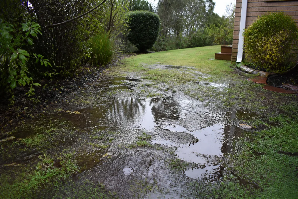 Terrain humide et boueux avec flaques, sol argileux et zone de ruissellement près d’une maison